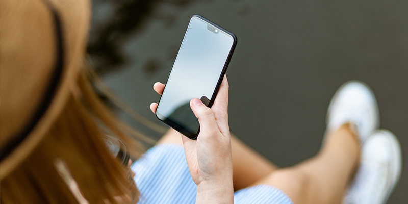 woman wearing brown brimmed hat, blue and white striped dress, and white shoes holds cell phone