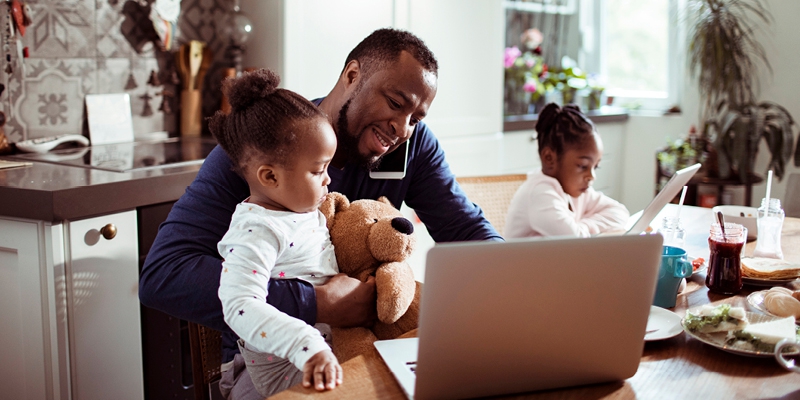 man at kitchen table using cell phone and laptop while holding one daughter and another daughter sits next to him