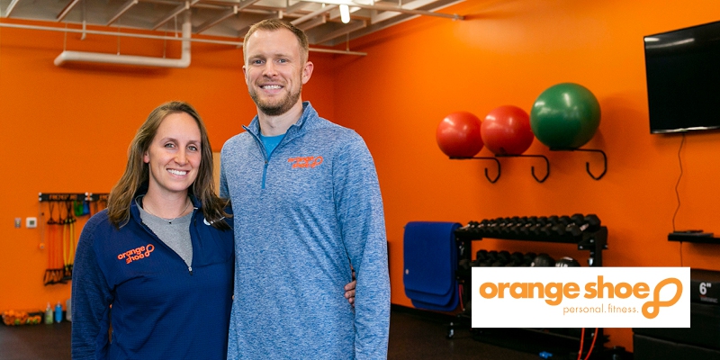 Emily and John McKiddy of Orange Shoe Personal Fitness smiling inside gym with orange walls with workout equipment behind them